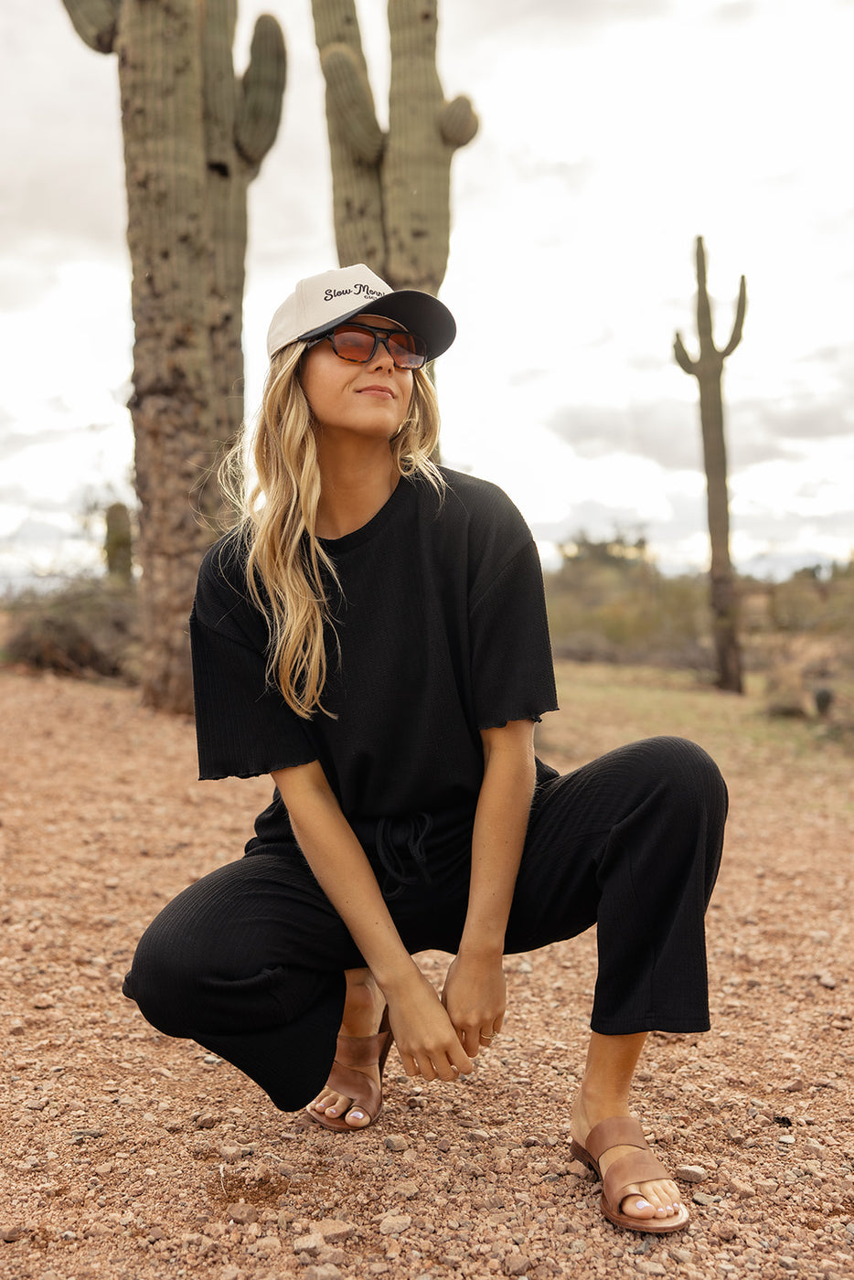 a woman in a hat and sunglasses squatting in the desert