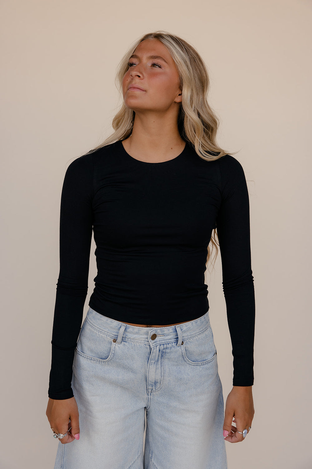 Woman wearing a black long-sleeve top and light blue jeans against a beige background
