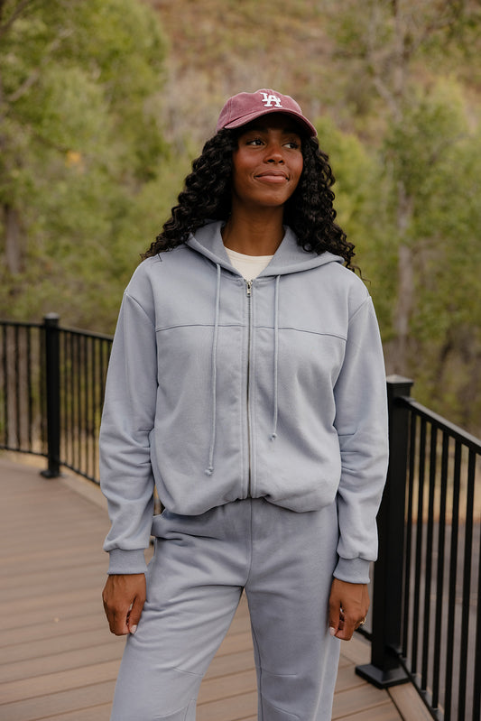 Person wearing a gray hoodie and pants set with a maroon cap outdoors.