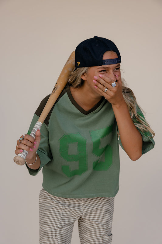 Person wearing a green jersey with the number 95, holding a baseball bat, and covering their mouth with their hand.