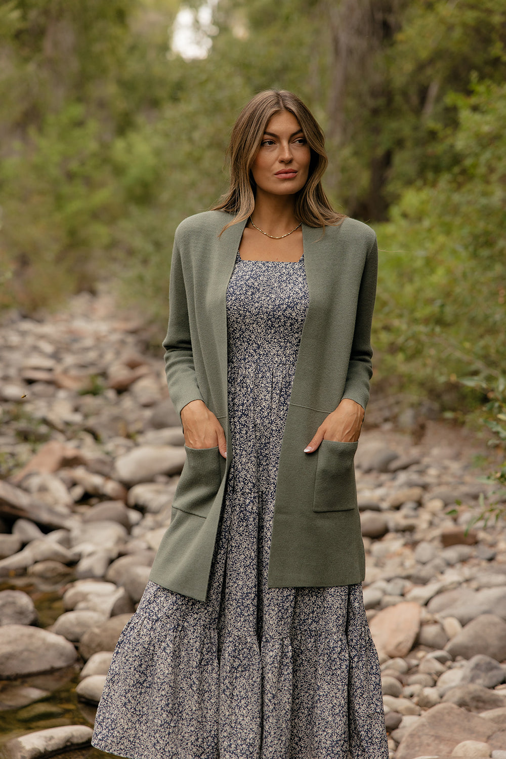 Woman wearing a green cardigan over a floral dress standing in a natural setting with rocks and trees.