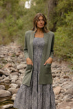 Woman wearing a green cardigan over a floral dress standing in a natural setting with rocks and trees.