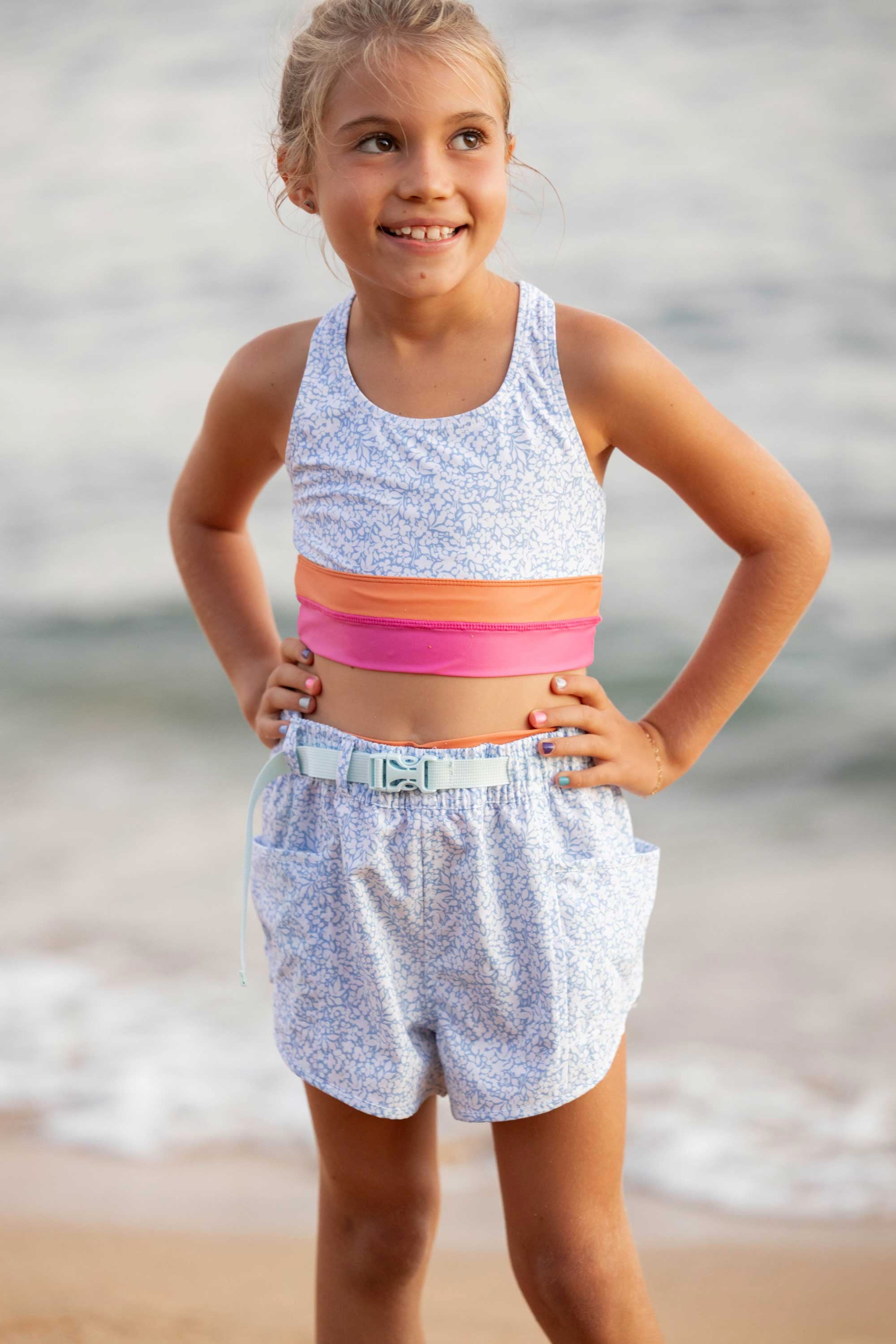 A young girl standing on a beach wearing a sleeveless top with blue and orange horizontal stripes and matching shorts with a floral pattern.