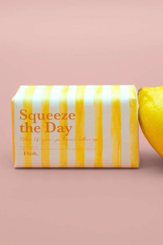 Bar of soap with yellow and white striped packaging labeled 'Squeeze the Day' next to a lemon on a pink background