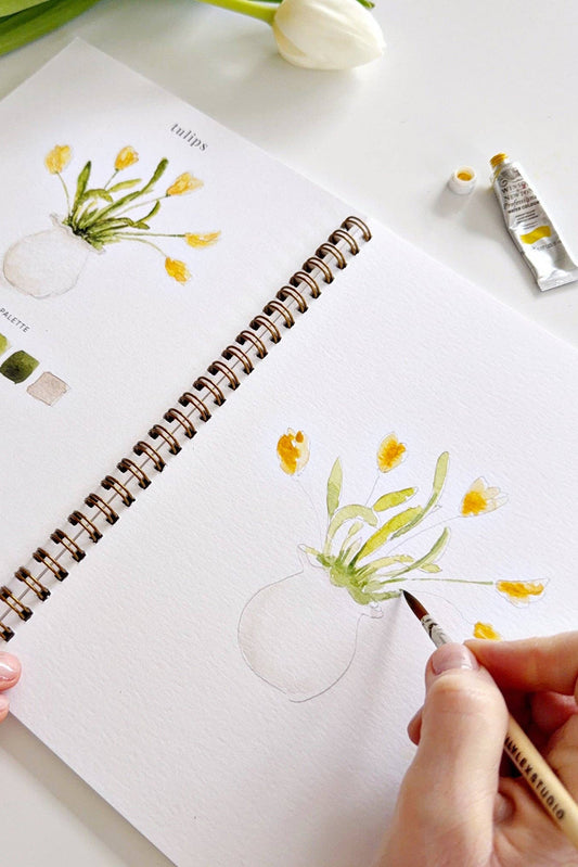 Person drawing flowers in a notebook with a paintbrush, surrounded by art supplies on a white surface.